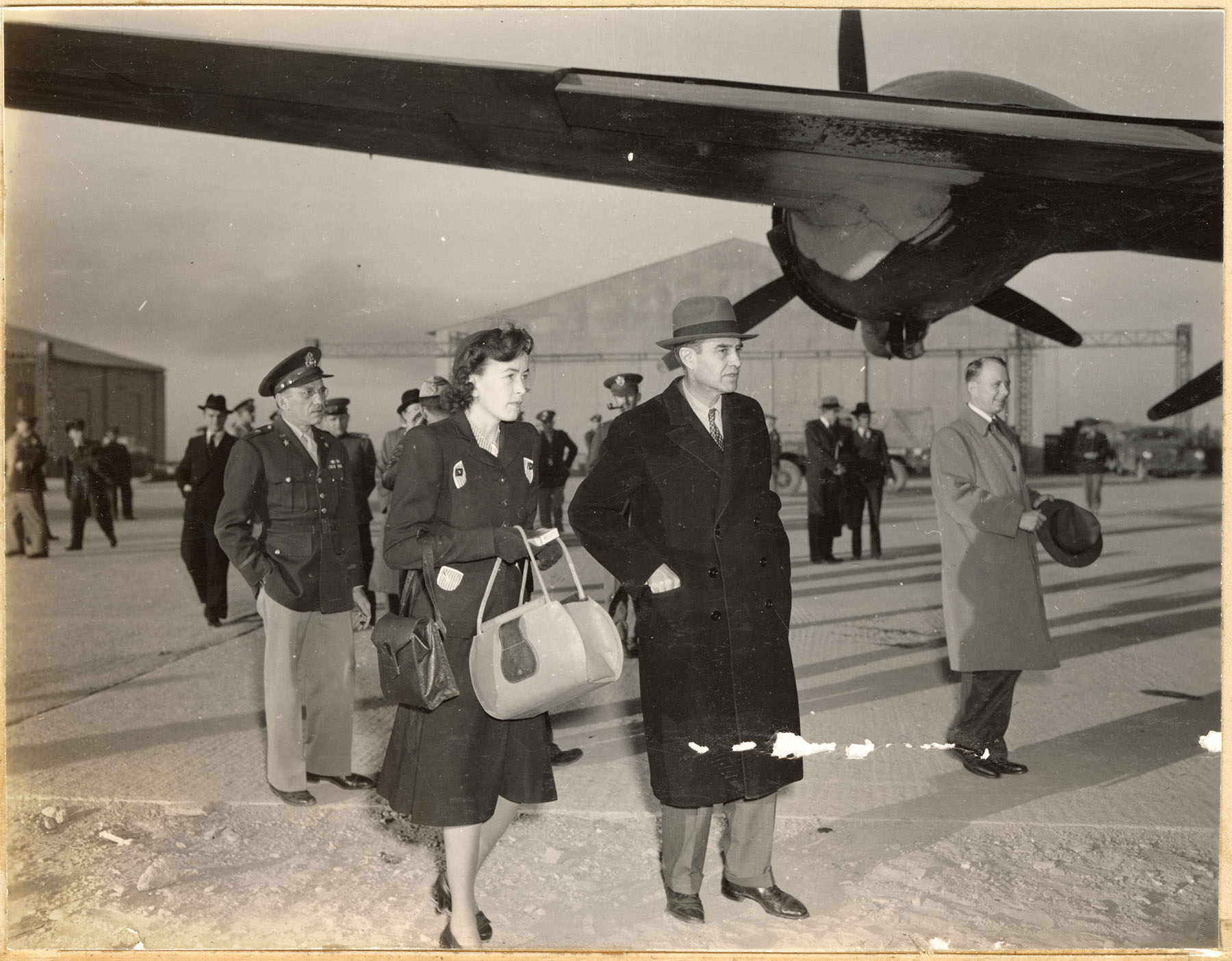 A civilian woman and man depart an airplane with army officers in backgroiund